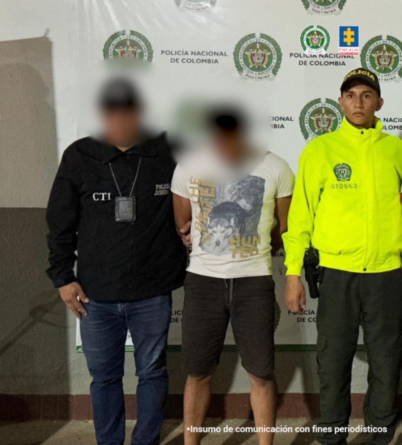 capturado de frente custodiado por un investigador del CTI de la Fiscalía y un policía. Detrás banner de la Policía Nacional.