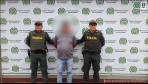 capturado de frente manos atrás esposado custodiado por dos policías. Detrás banner de la Policía Nacional.