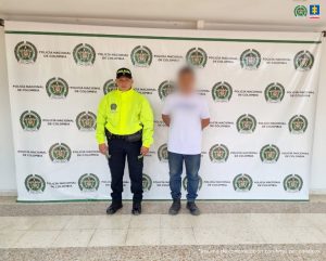 El imputado se encuentra custodiado por dos uniformados de la Policía Nacional. El capturado tiene una camisa rosada. Detrás se observa un backing de la Policía.