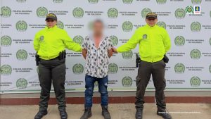  capturado de frente custodiado por dos policías.ñ Detrás banner de la Policía Nacional.
