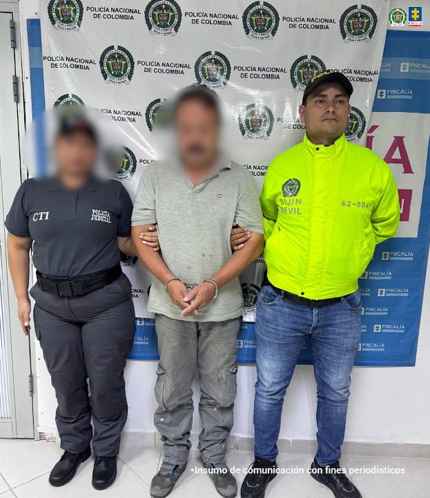 capturado de frente, custodiado por investigador del CTI de la Fiscalía y policía. Detrás banner de la Fiscalía.
