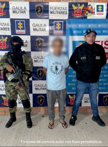 capturado de frente esposado manos atrás, custodiado por investigador de l CTI de la Fiscalía y un militar. Detrás banner de Fiscalía y Ejército.