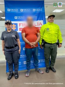 capturado de frente manos adelante esposado, custodiado por investigador del CTI de la Fiscalía y policía. Detrás banner de la Fiscalía.