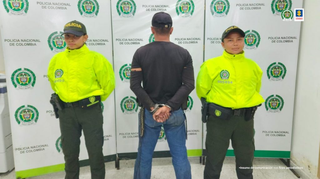 capturado de espaldascustodiado por dos policías. Detrás banner de la Policía Nacional.