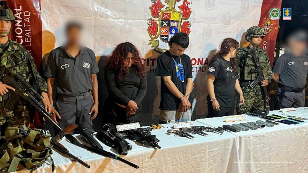 En la fotografía se aprecian de pie, dos de ellos con los brazos adelante y esposados a Juan Manuel Salgado Franco, Paola Andrea Sánchez Jiménez y Valentina Perilla Otálvaro. El hombre viste con camiseta de mangas cortas y color negro, así como Jean azul claro desgastado. Una de las mujeres usa el cabello suelto, rizado y viste con blusa de manga larga y pantalón negro. La otra mujer usa cabello largo, viste con camiseta de mangas cortas y estampado blando en mangas y pecho; también usa pantalón negro. En los extremos de ellos hay servidores del CTI de la Fiscalía y uniformados del Ejército Nacional. Delante de ellos hay una mesa con mantel blanco y sobre ella se aprecian armas de fuego largas y cortas, proveedores, celulares, munición, dinero en efectivo y un morral. Detrás de los imputados hay un pendón institucional del Ejército.