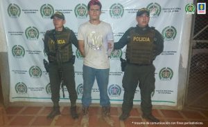  capturado de frente custodiado por dos policías. Detrás banner de la Policía Nacional.
