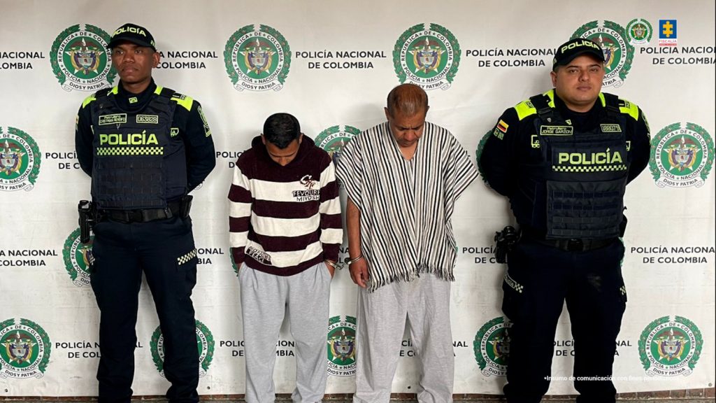 Fotografia en donde se pueden ver a los dos capturado en medio de dos funcionarios de la policia. Detras de ellos se ve un pendon de la Policia nacionl