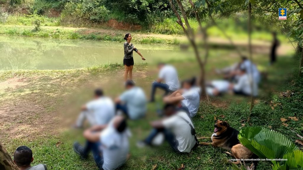Una mujer de pie dirige una actividad al aire libre frente a un grupo de personas sentadas en el pasto, cerca de un cuerpo de agua y rodeados de árboles. Las personas están escuchando mientras un perro descansa a un lado. La escena corresponde a una jornada de intervención y reflexión en un entorno natural.