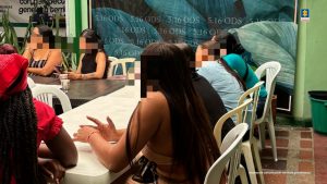 Varias personas sentadas alrededor de mesas plásticas participan en una actividad grupal en un espacio interior. Sus rostros están pixelados para proteger su identidad. El ambiente muestra un mural decorativo al fondo y sillas blancas alrededor. La escena corresponde a una jornada de intervención psicosocial relacionada con casos de violencia intrafamiliar.