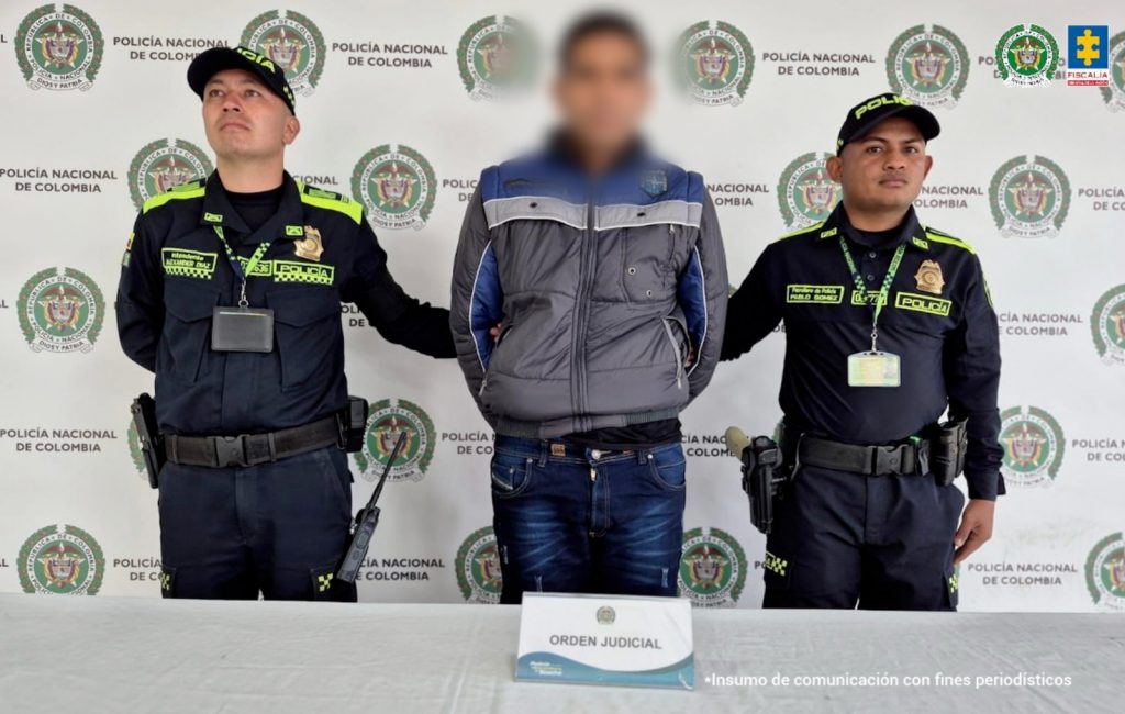 fotografia en donde se ve el capturado en medio de dos policias y detras de ellos un baking de su institución