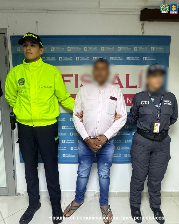 capturado de frente custodiado por investigador CTI de la Fiscalía y uniformado de la Policía Nacional. Detrás banner de la Fiscalía.
