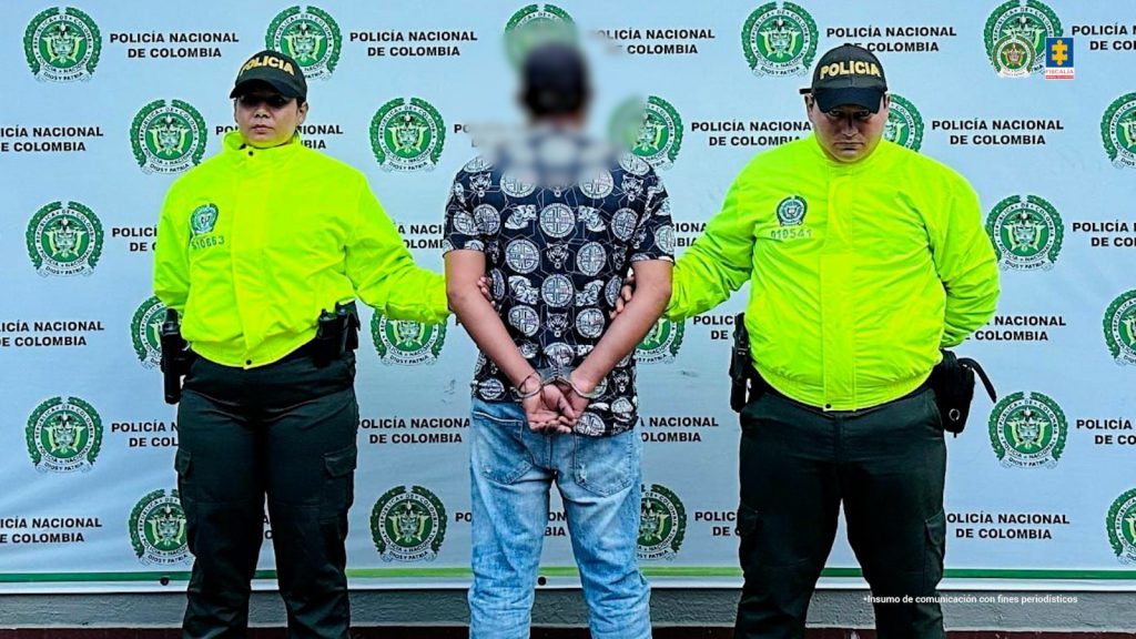 capturado de espaldas custodiado por dos policías. Detrás banner de la Policía Nacional.
