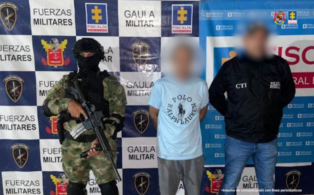 Capturado de frente custodiado por un militar y un investigador del CTI de la Fiscalía. Detrás banner de la Fiscalía y banner del Ejército Nacional.