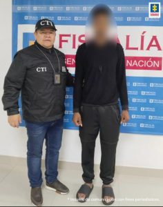 En la foto aparece de pie el procesado con buzo, pantalón negro y pantuflas negras. A su derecha hay un servidor del CTI de la Fiscalía y detrás de ellos hay un pendón institucional de la Fiscalía. 