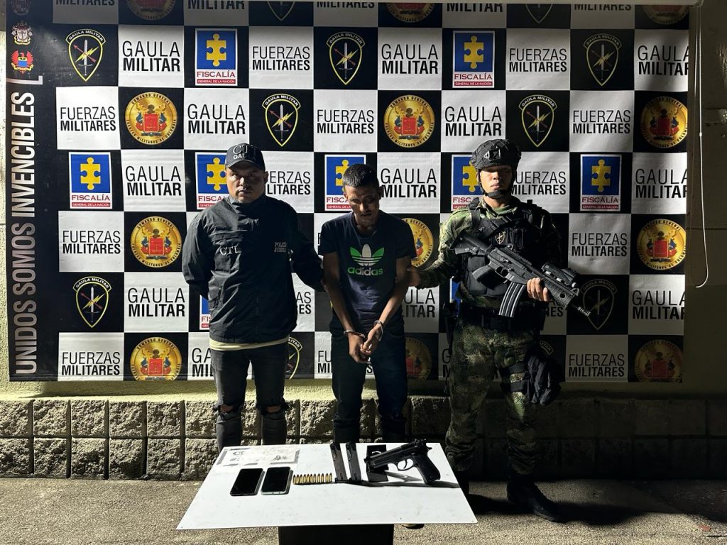 En la imagen aparece el procesado en medio un servidor del CTI y otro del Gaula del Ejército Nacional. Al frente hay una mesa con un arma de fuego tipo pistola.