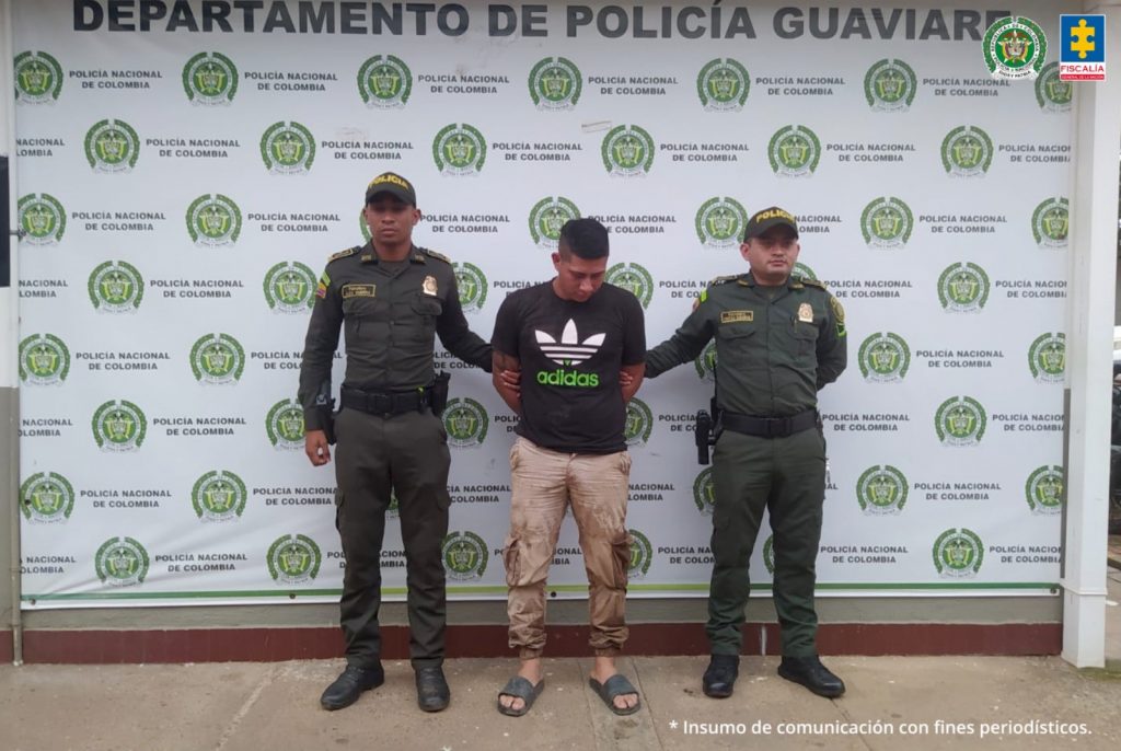 capturado de frente custodiado por dos policías. Detrás banner de la Policía Nacional.