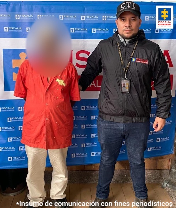 Se observa un hombre, de frente, capturado con efecto difuminado sobre su rostro. A su lado un funcionario del CTI. Detrás un pendón de la Fiscalía General de la Nación.