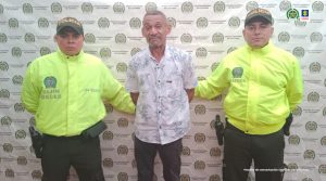En la fotografía aparece una persona capturada, acompañada de dos uniformados de la Policía Nacional. En la parte posterior un banner con logos de la entidad.