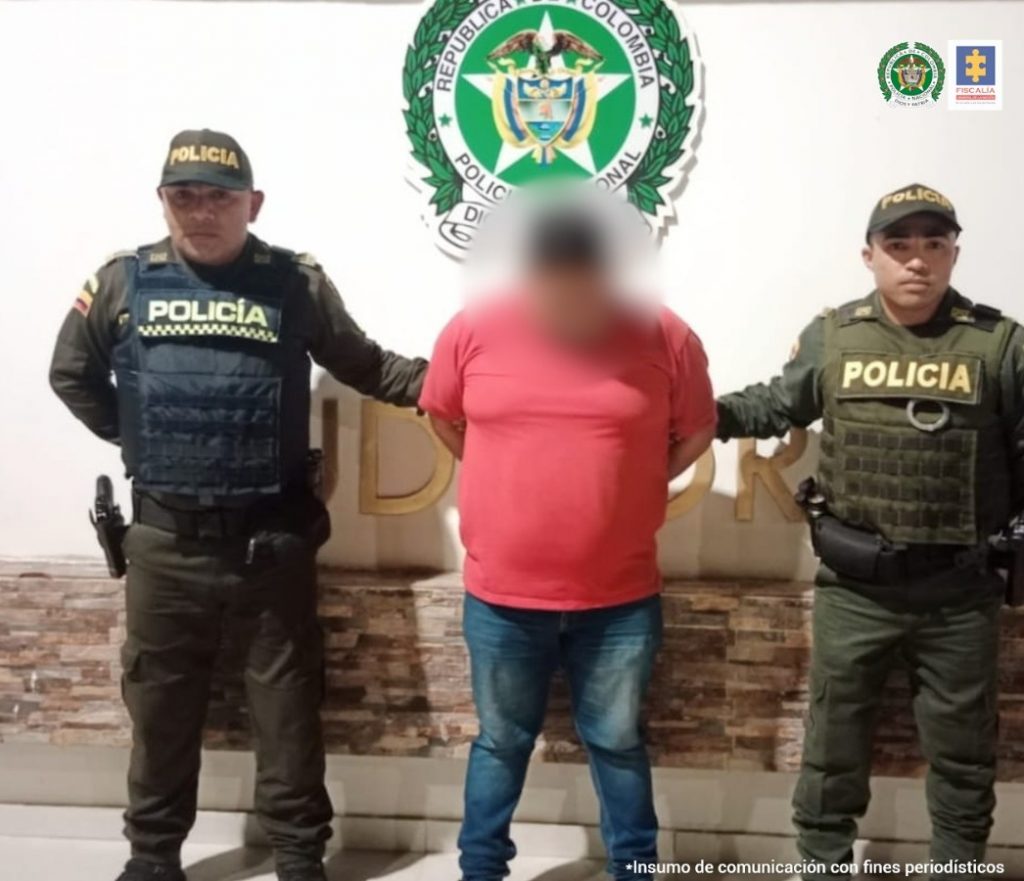 En la fotografía aparece un hombre capturado, acompañado de dos uniformados de la Policía Nacional. En la parte posterior un banner con logos de la entidad.