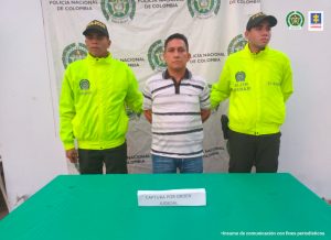 En la fotografía aparece una persona capturada, acompañada de dos uniformados de la Policía Nacional. En la parte posterior un banner con logos de la entidad.