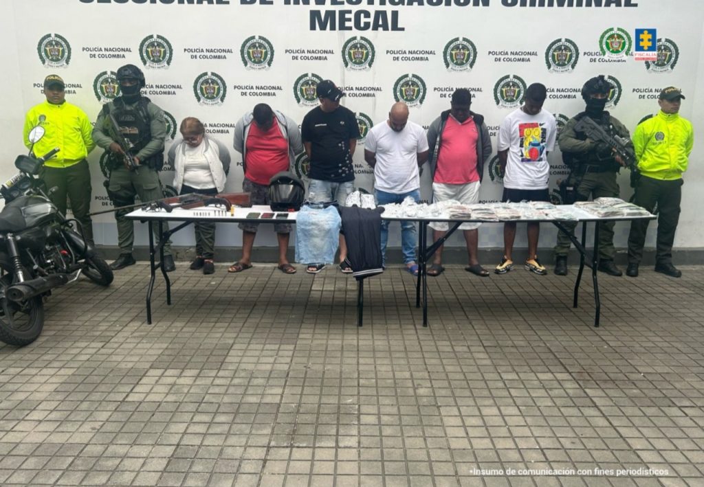 En la fotografía se aprecian de pie a los presuntos implicados. Entre ellos hay una mujer. Todos ellos tienen los brazos atrás y la cabeza agachada. A sus extremos hay uniformados de la Policía Nacional. Delante de ellos hay una mesa larga y sobre ella hay armas de fuego, munición, celulares, prendas de vestir, dinero en efectivo, bolsas plásticas con contenido blanco. Al extremo izquierdo de la fotografía se aprecia una motocicleta.