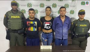 En la foto se aprecia de pie y con los brazos atrás a los tres presuntos implicados en la tentativa de hurto violento en Buga (Valle del Cauca. Uno de ellos viste camiseta de manga corta, negra con estampado de franjas verticales de colores a la altura del pecho y sobre ellas un control de videojuegos; así como pantalonera azul. La mujer viste con blusa negra pegada al cuerpo y arriba del ombligo y estampado el logo de ‘Fila’; asimismo, jean azul y zapatillas blancas. El otro hombre viste con camisa azul, de manga larga y de botones, jean azul y zapatillas blancas. A lado y lado de ellos hay uniformados de la Policía Nacional. Delante de ellos hay una mesa de color marrón y sobre ellas hay munición y un letrero que dice. ‘Porte ilegal de armas de fuego’. Detrás de ello hay un pendón institucional de la Policía.