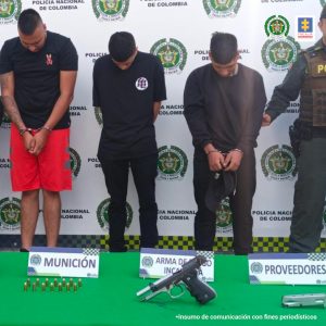 En la foto se aprecian a los tres hombres, con la cabeza agachada, dos de ellos con los brazos adelante y esposados, el otro con los brazos adelante. Uno de ellos viste buzo y pantalón negro y tiene una gorra negra en las manos; otro viste camisa de manga corta y pantalón negro y un tercero viste camiseta negra de manga corta negra y pantaloneta roja. Al lado izquierdo de ellos se aprecia un uniformado de la Policía Nacional. Delante de ellos hay una superficie verde y sobre ella hay un arma de fuego, un proveedor y munición; asimismo, hay tres letreros que dicen: ‘Munición’, ‘Arma de fuego incautada’ y ‘Proveedores’. Detrás de ellos hay un pendón institucional de la Policía.