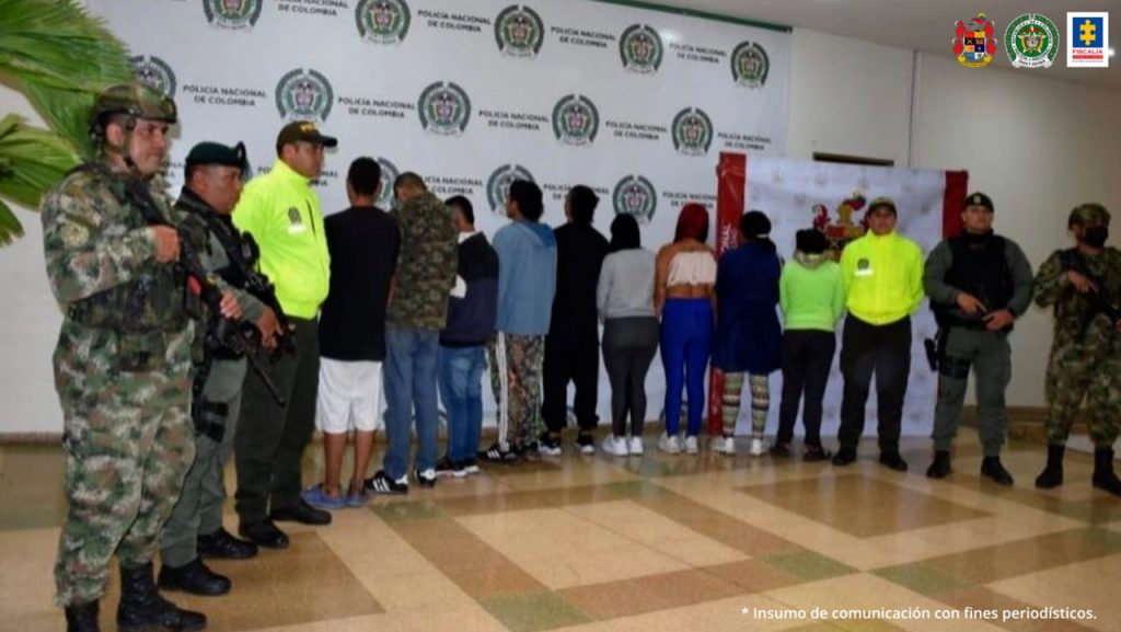 se observan 9 capturados y a cada extremo de la fila tres funcionarios de Policía Nacional y Ejército. Al fondo pendón de Policía nacional y de Ejército.