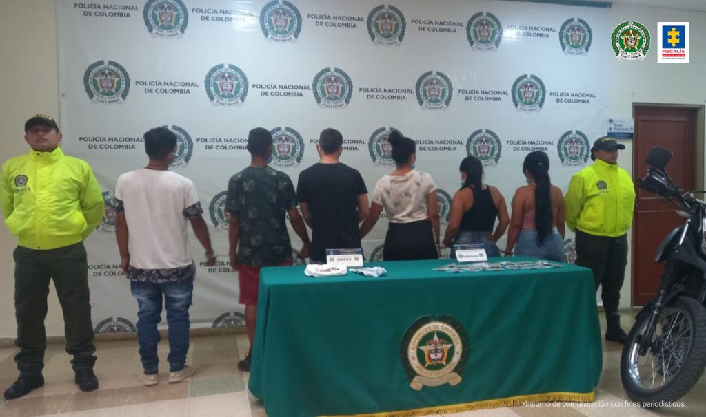 se observan seis capturados en fila y en cada lado un funcionario de Policía Nacional En un extremo una motocicleta En el centro una mesa con elementos incautados en operativos al fondo backing de la Policía Nacional