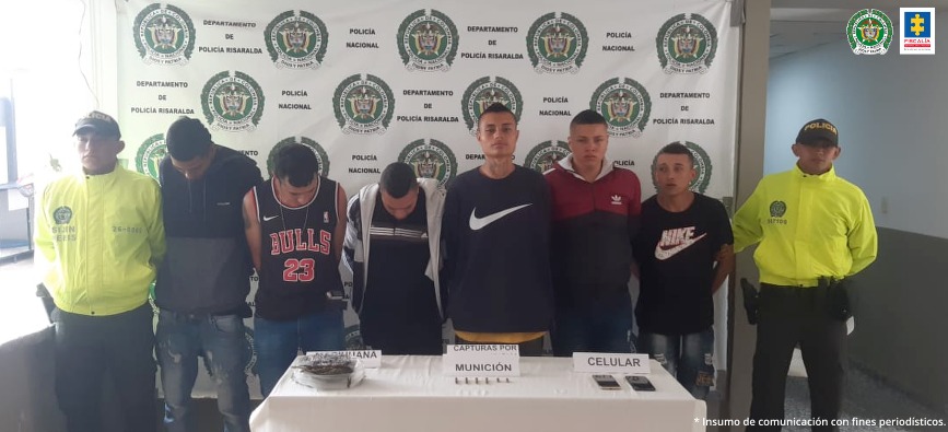 Se observan 6 capturados en fila y en cada lado un funcionario de la Policía Nacional. En el fondo un backing de la Policía Nacional. Al frente de los capturados en una mesa se observan elementos incautados durante diligencias de allanamiento.