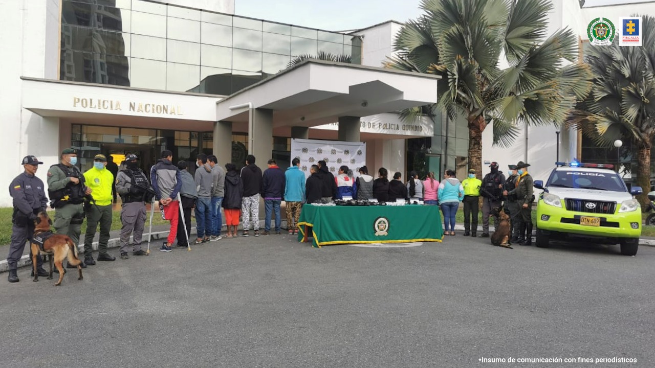 Se observan 17 personas capturadas, todas de espaldas y en fila. En ambos costados personal de la Policía y del CTI de la Fiscalía General de la Nación. Igualmente se ven dos caninos que acompañaron los operativos. En el centro una mesa con mantel verde en la que se encuentra el material incautado durante las capturas.