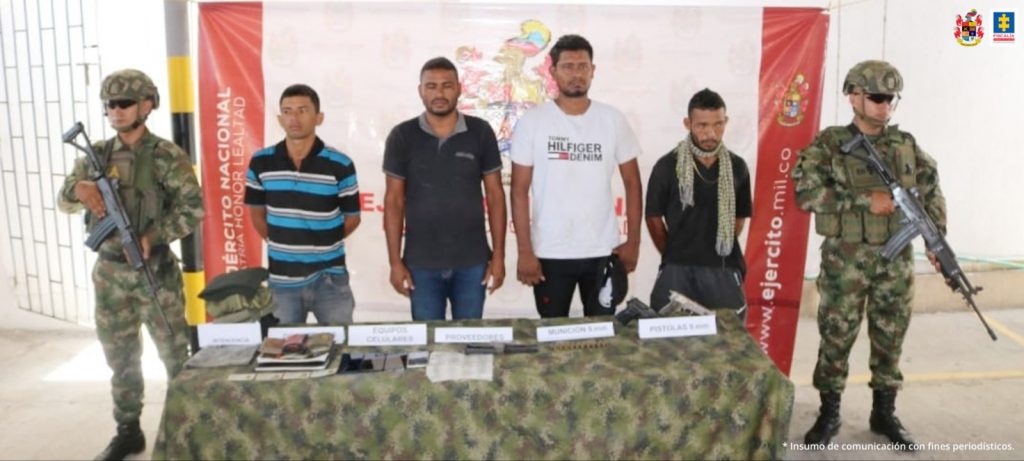 En la fotografía aparecen cuatro personas capturadas, acompañados de dos uniformados del Ejército Nacional. En la parte posterior un banner con logos de la entidad.