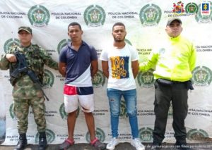 En la fotografía aparecen dos hombres capturados, acompañados de uniformados del Ejército y la Policía Nacional. En la parte posterior un banner con logos de la entidad.