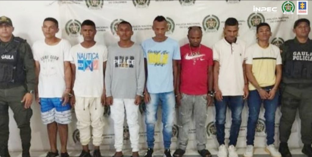 En la fotografía aparecen siete personas notificadas de la orden de captura en la Cárcel de Riohacha (La Guajira), acompañados de uniformados de la Policía Nacional. En la parte posterior un banner con logos de la entidad.