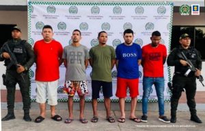 En la fotografía aparecen cinco personas capturadas, acompañados de un uniformado de la Policía Nacional. En la parte posterior un banner con el logo de la entidad.