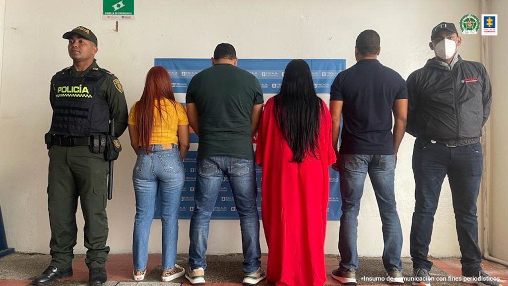 En la fotografía aparecen cinco personas capturadas dos mujeres capturadas, junto a dos uniformados de la Policía Nacional. En la parte posterior un banner de la Fiscalía General de la Nación.