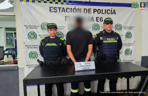 En la fotografía se aprecia de pie y de espaldas a la cámara al presunto responsable de tocar y abusar sexualmente de su propia hija. Viste camisa negra de magna corta, panalón negro con rayas en las pantorrillas verde y blanco fluorscentes. A los extremos de él hay uniformados de la Policía Nacional. En la parte delantera hay una superficie marrón y sobre ella hay un letrero que dice: ‘Capturado con orden judicial’. En la parte posterior hay un pendón institucional de la Policía.