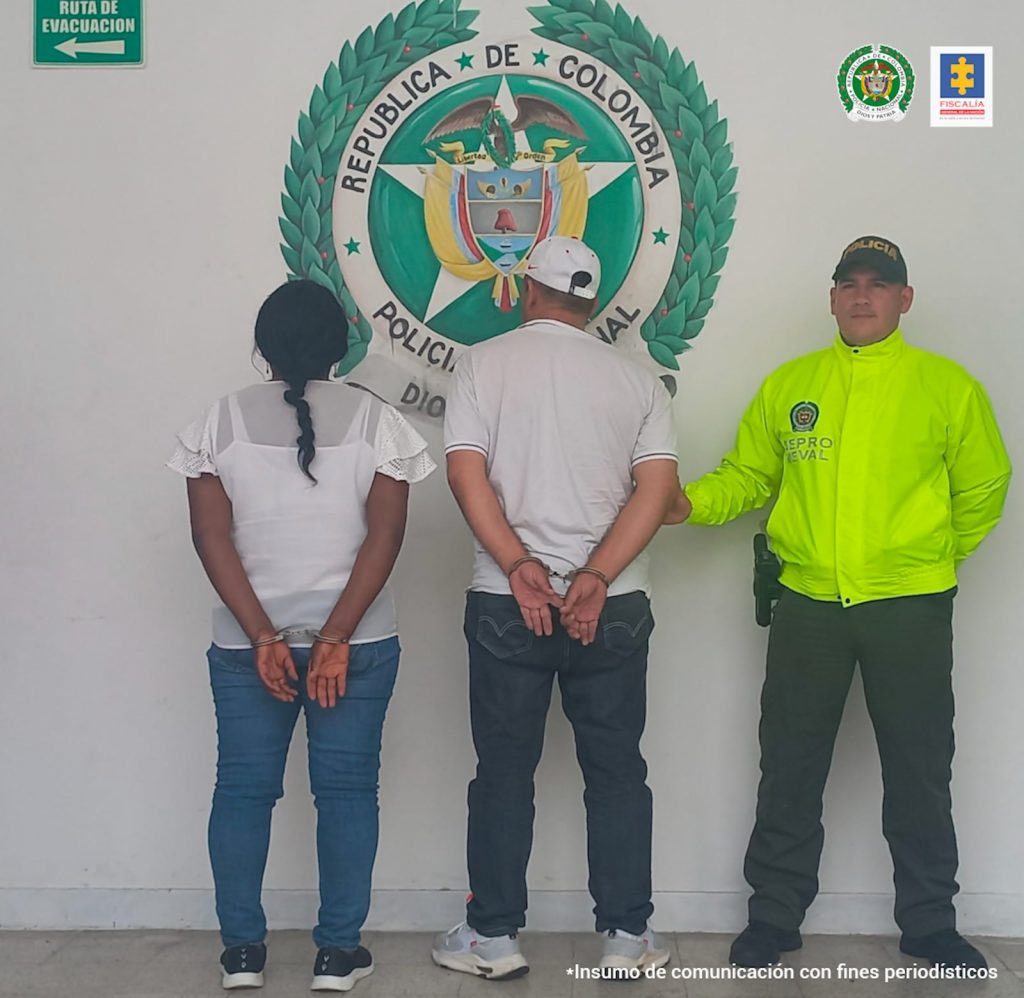 En la fotografía están los dos investigados de espalda con un uniformado de la Policía Nacional y un pendón de esa institución. 