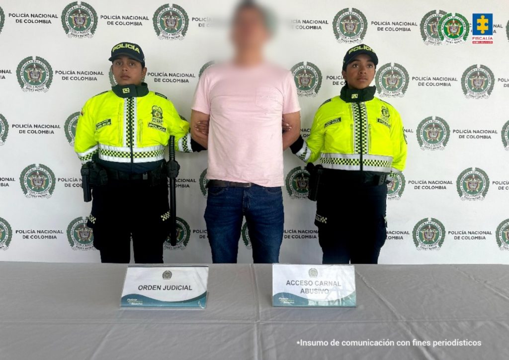 fotografia del capturado con el rostro borroso en medios de dos agentes de policia, frente a ellos una mesa con dos carteles que dicen: oden judicial y el otro acceso carnal abusivo. detras aparece un baking de la policia