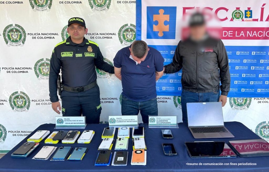 En la foto aparece el capturado de frente con buso azul oscuro y custodiado por un servidor del Cuerpo Técnico de Investigación (CTI) y un uniformado de la Policía Nacional. En la foto aparece de fondo un backing de la Fiscalía y Policía Nacional y los equipos electrónicos incautados.
