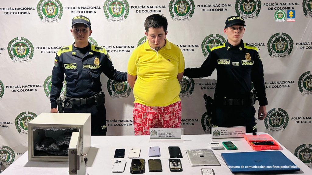 En la foto aparece el capturado con buso de color amarillo de frente y custodiado por dos uniformados de la Policía Nacional. En la foto aparece de fondo un backing de la Policía Nacional y los equipos electrónicos incautados.