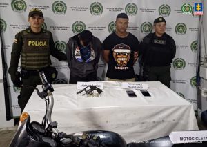 En la imagen aparecen dos personas de suéter negro detenidas entre dos uniformados de la Policía frente a una mesa con dos celulares y un arma de fuego.Boletín 