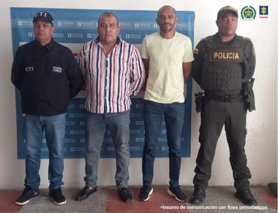 En la fotografía aparecen dos hombres capturados, acompañados de un servidor del CTI y un uniformado de la Policía Nacional. En la parte posterior un banner con los logos de la Fiscalía y la Policía.