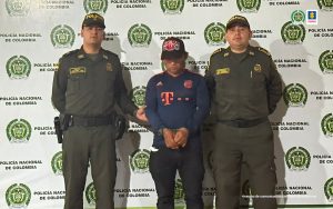 capturado de frente con gorra manos adelante esposado, custodiado por dos policías. Detrás banner de la Policía Nacional.
