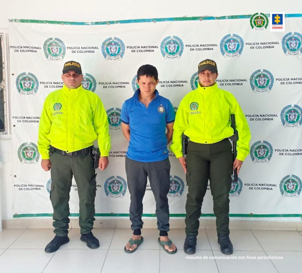 En la fotografía aparece un capturado, junto a personal de Policía Nacional. En la parte posterior de la imagen se ven banners de Policía Nacional y la Fiscalía General de la Nación.  