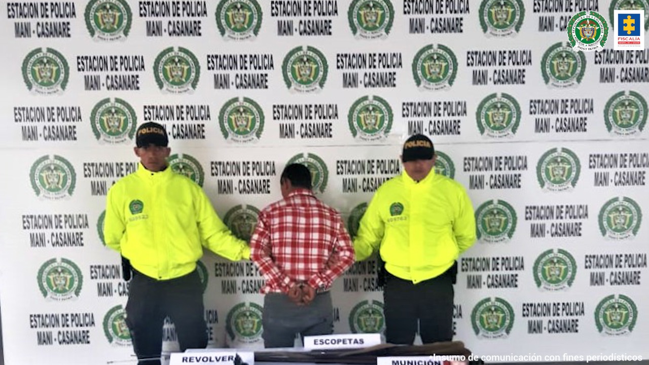 El captuado se encuentra de espaldas a la mesa donde stán las 3 armas que se le incautaron, y está protegido por dos policías uniformados.