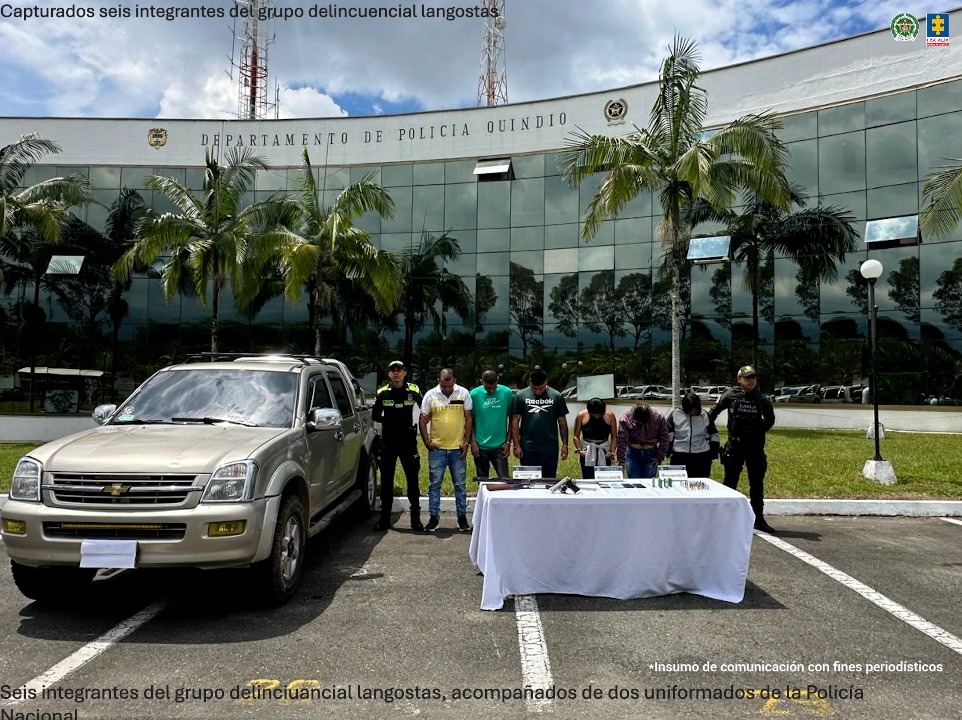 En primer plano hay una camioneta color beige y una mesa cubierta con un mantel blanco donde reposan armas de fuego, municiones y otros elementos incautados. Frente a la mesa están seis personas capturadas, acompañadas por dos uniformados de la Policía Nacional.