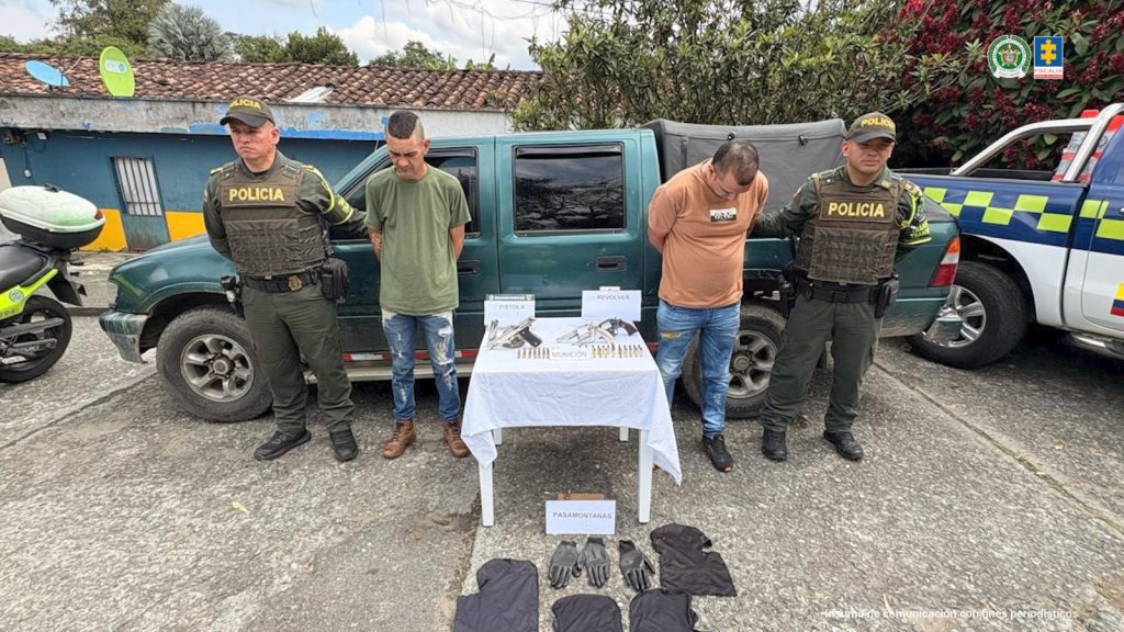 en la imagen se ven los dos capturados en medio de dos agentes de policia, delante de ellos se ve una mesa con las armas, municiones y accesorios incautados, atras de ellos se ve una camioneta verde