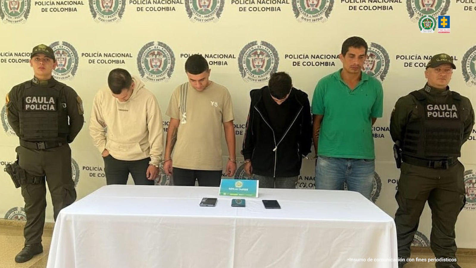 En la imagen aparecen cuatro hombres capturados, esposados y custodiados por dos uniformados del GAULA de la Policía Nacional. Los capturados están de pie frente a un fondo institucional con logos de la Policía Nacional y la Fiscalía General de la Nación. Sobre una mesa blanca, ubicada en primer plano, se observan tres teléfonos celulares incautados.
