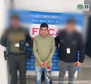 Se visualiza al capturado junto a un uniformado de la Policía y un investigador del CTI. Detrás de ellos el banner que identifica a la Fiscalía General de la Nación.